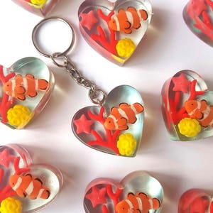 Könnte beinhalten: Herzförmige Schlüsselanhänger mit einem Design aus klarem Harz mit einem orange-weiß gestreiften Clownfisch, roter Koralle, gelber Seeanemone und rosa Seesternen. Der Schlüsselanhänger hat einen silbernen Metallring und eine Kette.