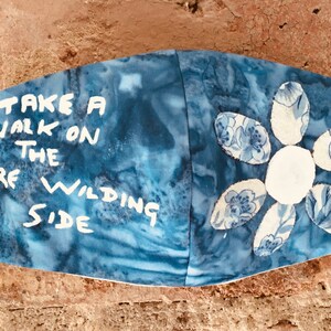 Puede incluir: Máscara facial de tela azul con texto blanco que dice "Take a walk on the wild side" y una flor blanca con pétalos azules.