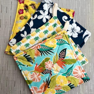 May include: Set of three colorful pot holders with tropical patterns. The pot holders feature pineapples, hibiscus flowers, and palm leaves.