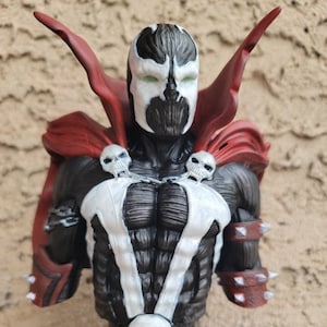May include: A black and white bust of the comic book character Spawn. The bust is made of plastic and features a detailed sculpt of Spawn's head, chest, and arms. Spawn is wearing his signature red cape and has a skull on his chest.