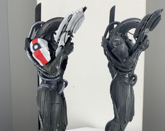 Op de afbeelding: Twee 3D-geprinte figuren van een wezen met een wapen, gemaakt van zwart en grijs. De figuur links heeft een wit en rood ontwerp op zijn arm. Beide figuren staan op zwarte ronde bases.