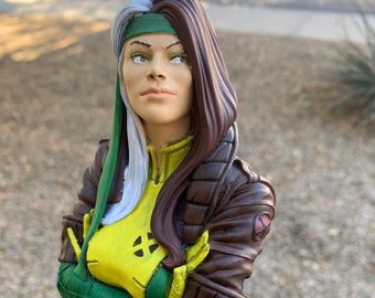 May include: A green, yellow, and brown bust of a female superhero with white hair. The bust is posed with her arms crossed and she is wearing a green and yellow costume with a brown jacket.