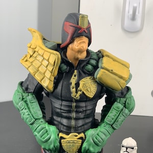 May include: A green, black, and yellow action figure bust of Judge Dredd, a fictional character from the 2000 AD comic book series. The bust is on a brick-like base and is standing next to a white and red action figure.