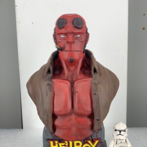 May include: A red and brown bust of Hellboy, a fictional superhero, with a cigar in his mouth. The bust is on a black base with the word "HELLBOY" in yellow letters. A white action figure of a Stormtrooper stands in front of the bust.