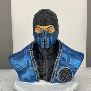 May include: A blue and black bust of Sub-Zero from the Mortal Kombat video game series. The bust is made of plastic and features a detailed sculpt of the character's face and costume. The bust is sitting on a white base with the Mortal Kombat logo.