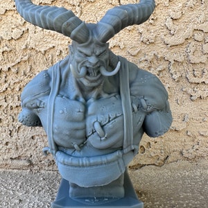 May include: A gray 3D printed bust of a demon with large horns, a fierce expression, and a stitched-up chest. The bust is mounted on a gray base.