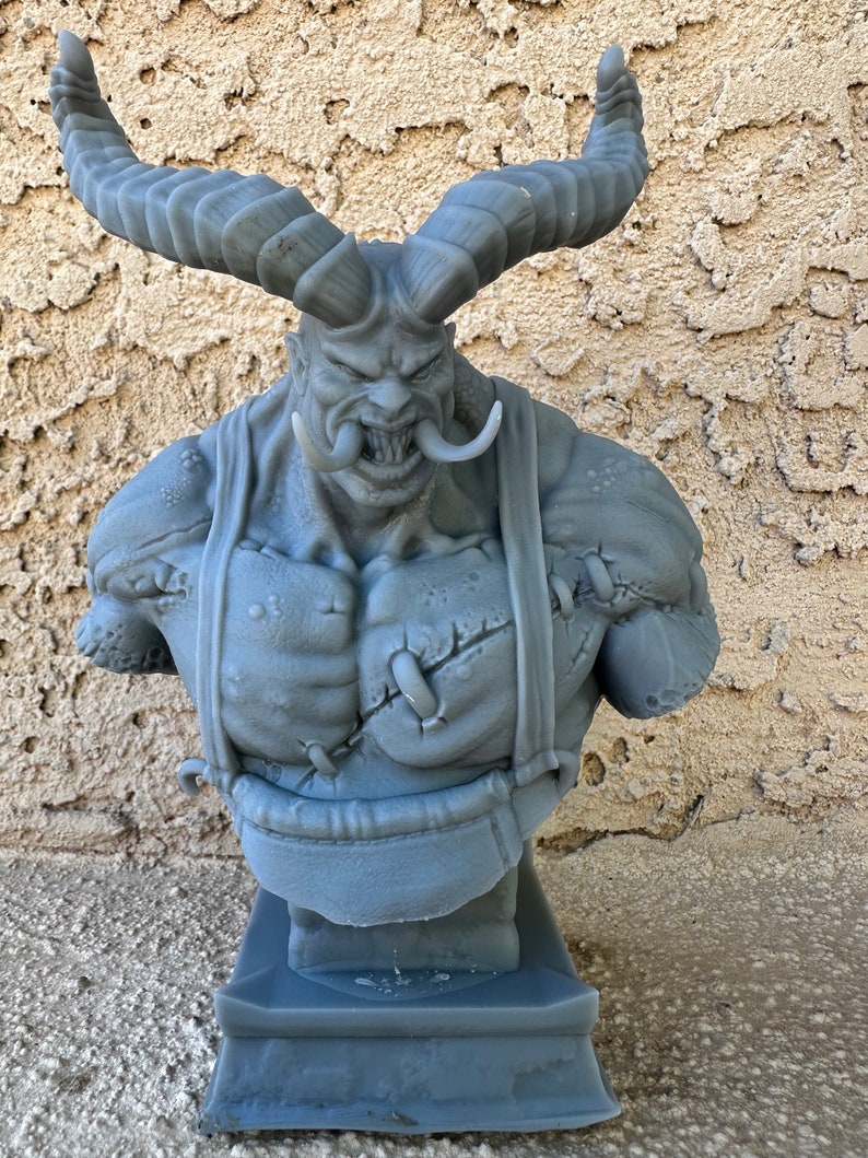 May include: A gray 3D printed bust of a demon with large horns, a fierce expression, and a stitched-up chest. The bust is mounted on a gray base.