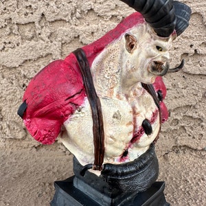 May include: A bust of a monstrous creature with a large horn, red and white skin, and a black leather strap across its chest. The bust is on a black base.