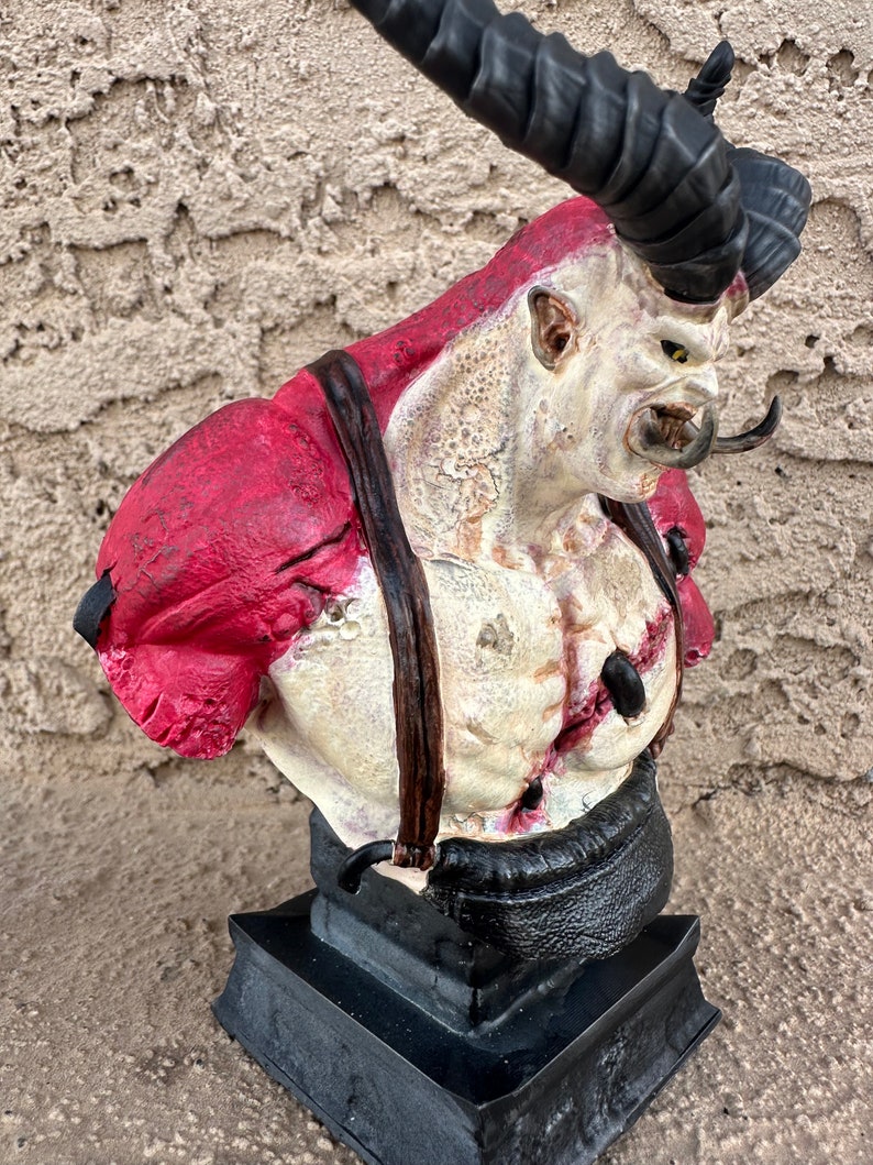 May include: A bust of a monstrous creature with a large horn, red and white skin, and a black leather strap across its chest. The bust is on a black base.