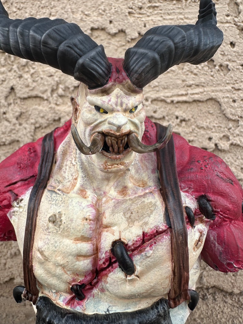 May include: A red and white painted bust of a demon with large black horns, a wide mouth, and a stitched-up chest. The demon has a menacing expression.