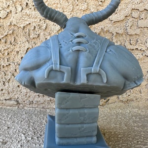 May include: A gray, 3D printed sculpture of a creature with horns, a stitched-up body, and a base made of stacked blocks. The sculpture is made of a smooth, gray material.