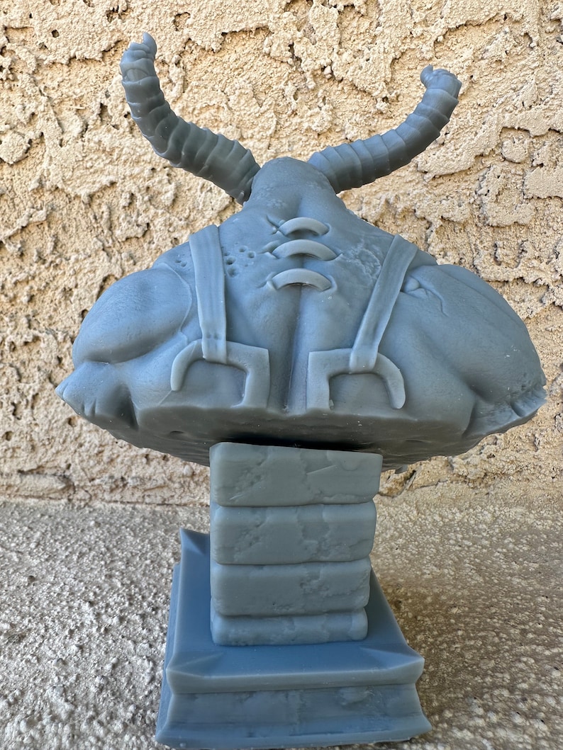 May include: A gray, 3D printed sculpture of a creature with horns, a stitched-up body, and a base made of stacked blocks. The sculpture is made of a smooth, gray material.