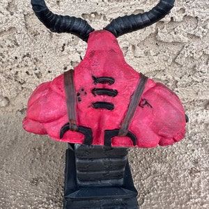May include: A red and black demon bust figurine with horns. The demon is wearing a leather harness with straps and buckles. The bust is sitting on a black base.