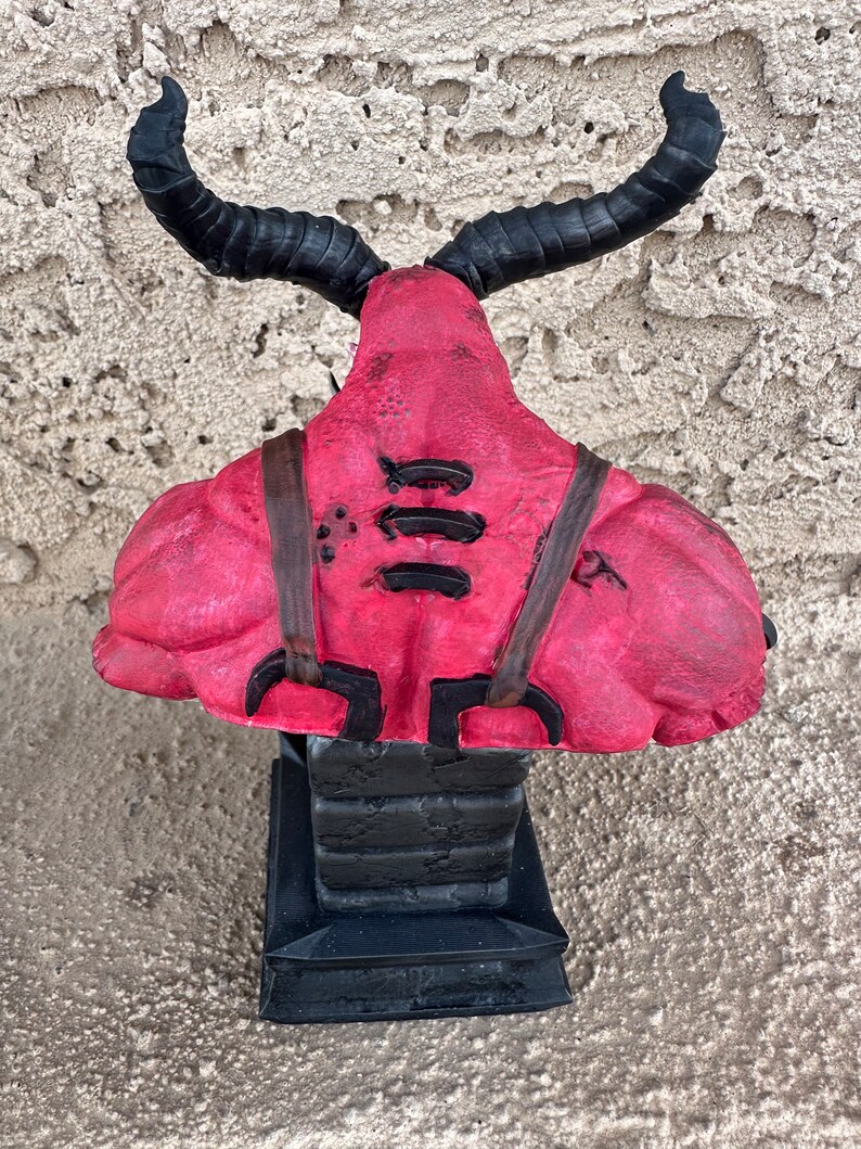 May include: A red and black demon bust figurine with horns. The demon is wearing a leather harness with straps and buckles. The bust is sitting on a black base.