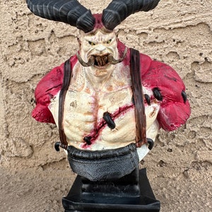 May include: A red and black painted bust of a horned, muscular figure with a large mustache and a menacing expression. The figure is wearing a red shirt with straps and a black belt. The bust is mounted on a black base.