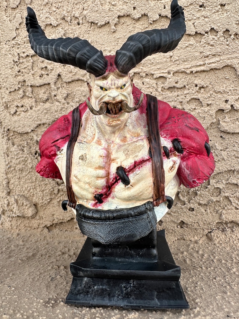 May include: A red and black painted bust of a horned, muscular figure with a large mustache and a menacing expression. The figure is wearing a red shirt with straps and a black belt. The bust is mounted on a black base.