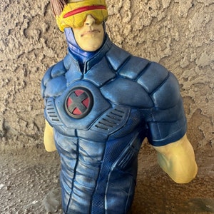 May include: A blue and yellow bust of a superhero with a red X on the chest. The superhero is wearing a yellow visor with red accents.