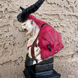 May include: A red and white demon bust with black horns and a black base. The demon has a fierce expression and is wearing a red garment.