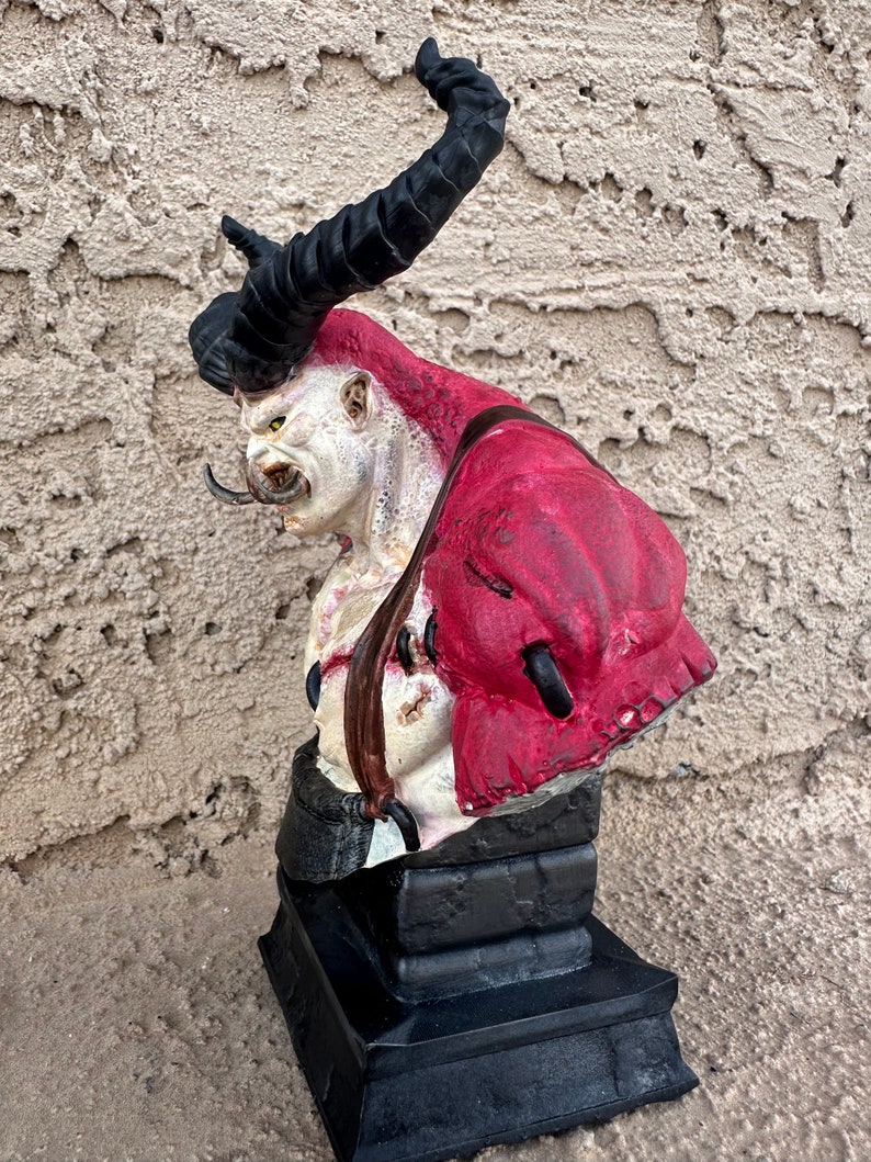 May include: A red and white demon bust with black horns and a black base. The demon has a fierce expression and is wearing a red garment.