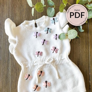 Puede incluir: Un body de bebé blanco tejido con bordado de libélulas en colores pastel. El body tiene una cintura con lazo, mangas con volantes y un cuello redondo. La imagen incluye un icono PDF digital.