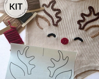 May include: A beige knit sweater featuring a reindeer face design with dark brown antlers, black eyes, and a red nose. The kit includes spools of brown, black, and red yarn, along with a white paper template showing the reindeer outline and the text KIT.