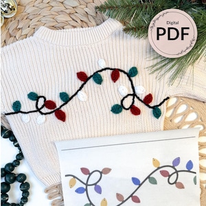 Puede incluir: Suéter color crema con un diseño festivo de luces navideñas en rojo, verde y blanco. Se incluye un patrón PDF digital, junto con una página impresa que muestra el diseño. El suéter está sobre una superficie tejida.