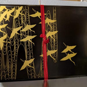 May include: A rectangular black lacquer box featuring a gold-colored graphic of flying cranes and swirling wave patterns. A thin red ribbon is tied vertically around the center of the box, securing it with a small knot at the front surface.