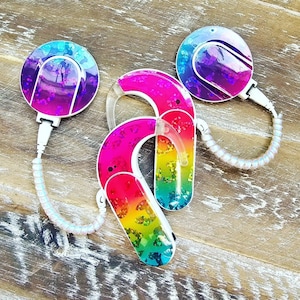 May include: Two rainbow-colored hearing aids with a sparkly finish and a white cord. The hearing aids are connected to a small, round, sparkly purple and pink device with a white cord.