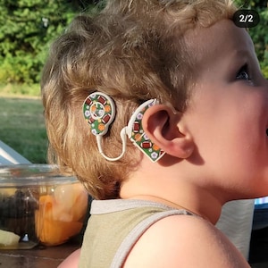 Puede incluir: Un niño pequeño usando un audífono con un diseño deportivo verde, blanco y naranja. El audífono está detrás de la oreja derecha del niño.