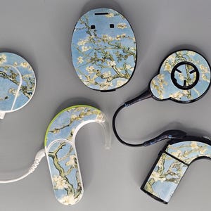 Puede incluir: Un conjunto de cuatro dispositivos de audífonos con un patrón floral. Los dispositivos son de varias formas y tamaños, incluyendo un dispositivo redondo, un dispositivo rectangular pequeño y dos dispositivos curvos. El patrón floral es un diseño azul claro y blanco con una rama de flores blancas.