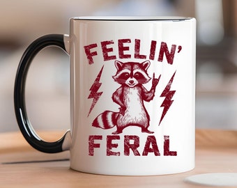 May include: White ceramic coffee mug with a black handle. The mug features a red graphic of a raccoon with lightning bolts and the text "Feelin' Feral".