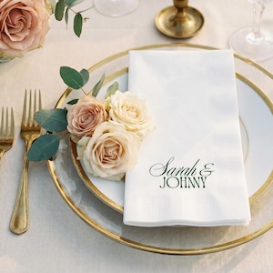 Pode incluir: Elegante mesa com pratos dourados, talheres dourados e taças de cristal. Uma toalha de mesa branca com "Sarah & Johnny" impressa em verde é colocada em um prato, adornada com rosas e vegetação. Uma vela acesa em um castiçal dourado.