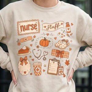 Op de afbeelding: Een beige sweater met ronde hals en een grafische print met de tekst "nurse stuff" omringd door illustraties van een pompoen, een stethoscoop, een klembord, een koffiebeker, een pleister, een stuk taart en herfstbladeren.
