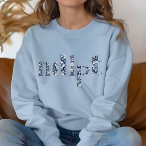 May include: A light blue sweatshirt with a floral pattern spelling out the word "ndipt".