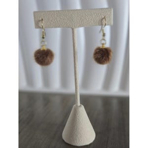 May include: Brown faux fur pom-pom earrings with gold-colored hooks and clear beads. The earrings are displayed on a beige jewelry stand against a white backdrop. The earrings are a simple, yet elegant accessory.