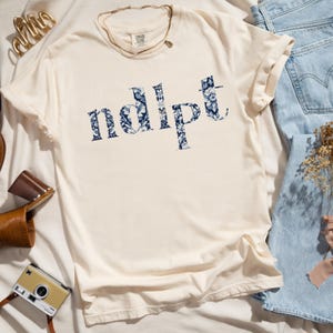 May include: A cream colored t-shirt with a blue floral print that spells out the word "ndipt".