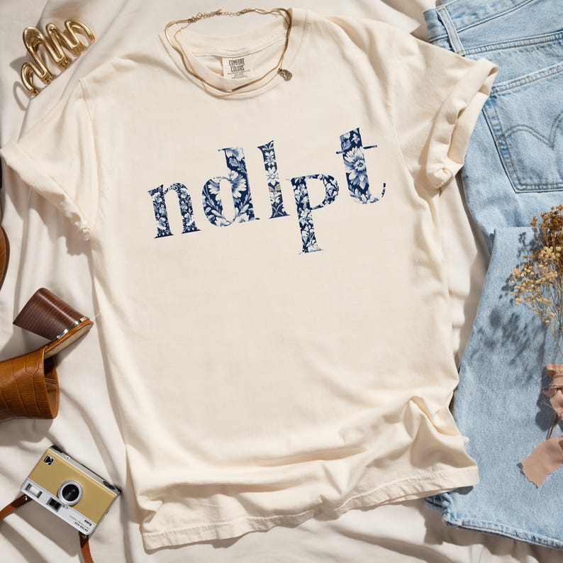 May include: A cream colored t-shirt with a blue floral print that spells out the word "ndipt".
