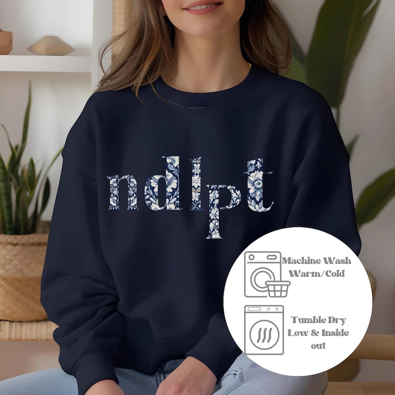 May include: Navy blue sweatshirt with the word "ndipt" printed in white letters with a floral pattern. The sweatshirt has a crew neck and long sleeves. The image also includes care instructions for the sweatshirt, which include machine washing in warm or cold water and tumble drying on low heat.