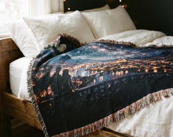 May include: A woven throw blanket featuring a dark night scene with a starry sky, a glowing city skyline, and a bridge over water. The blanket has a fringed edge and displays a silhouette of two figures standing on the left side of the design.