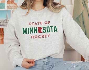 Pode incluir: Uma camisa de moletom cinza com o texto "STATE OF MINNESOTA HOCKEY" em letras vermelhas e verdes. O estado de Minnesota é retratado em vermelho dentro do texto.