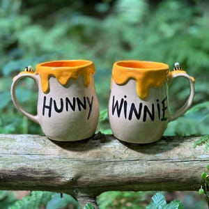 Könnte beinhalten: Zwei Keramikbecher mit beigefarbenem Boden und einem gelben, honigartigen Tropfdesign am Rand. Auf einem Becher steht "HUNNY" und auf dem anderen "WINNIE" in schwarzen Buchstaben. Jeder Becher hat eine Biene am Henkel.