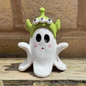 May include: A white ceramic ghost figurine wearing a lime green alien hat with three eyes and an antenna. The ghost has two large black oval eyes, a small red circular mouth, and two pink circular cheeks, with arms extended outward to the sides.
