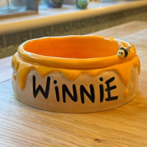 May include: A ceramic bowl with an orange interior and a yellow exterior designed to look like dripping honey. The word "WINNIE" is written in black letters on the bowl. A small bee figurine is attached to the rim of the bowl.