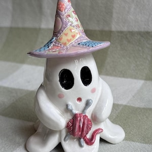 Può includere: Una statuetta di fantasma in ceramica bianca che indossa un cappello conico con motivo patchwork nei toni del rosa, giallo e blu. Il fantasma ha occhi ovali neri, piccole guance rosa e tiene in mano un gomitolo di lana rosa scuro con due ferri da maglia grigi.