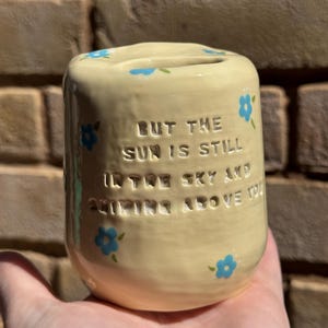 May include: A light yellow ceramic bank with blue floral accents. The bank has a slot on top and features the quote, "BUT THE SUN IS STILL IN THE SKY AND SHINING ABOVE YOU."