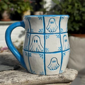 Peut inclure: Une tasse en céramique avec une base blanche et un motif à carreaux bleus. Chaque carré du quadrillage présente une petite figurine de fantôme peinte à la main. La tasse possède une anse bleue unie et une finition brillante avec des motifs floraux.