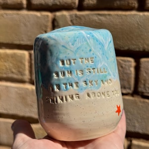 May include: A ceramic tumbler with a light blue and white sky design on top and a sandy beige base. The tumbler has the text "But the sun is still in the sky and shining above you" and a small red star.