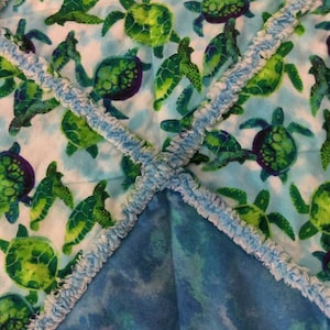 May include: A soft blue and white baby blanket with a green and purple sea turtle pattern. The blanket has a blue and white ruffled trim.
