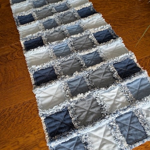 May include: A rectangular rag quilt table runner constructed from various shades of blue denim and light blue fabric squares. Each square features a frayed edge and an X-shaped stitch pattern across the center, creating a textured surface.