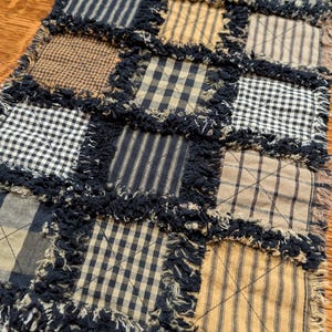 May include: A black and tan patchwork table runner with frayed edges. The runner is made of various plaid and striped fabrics.